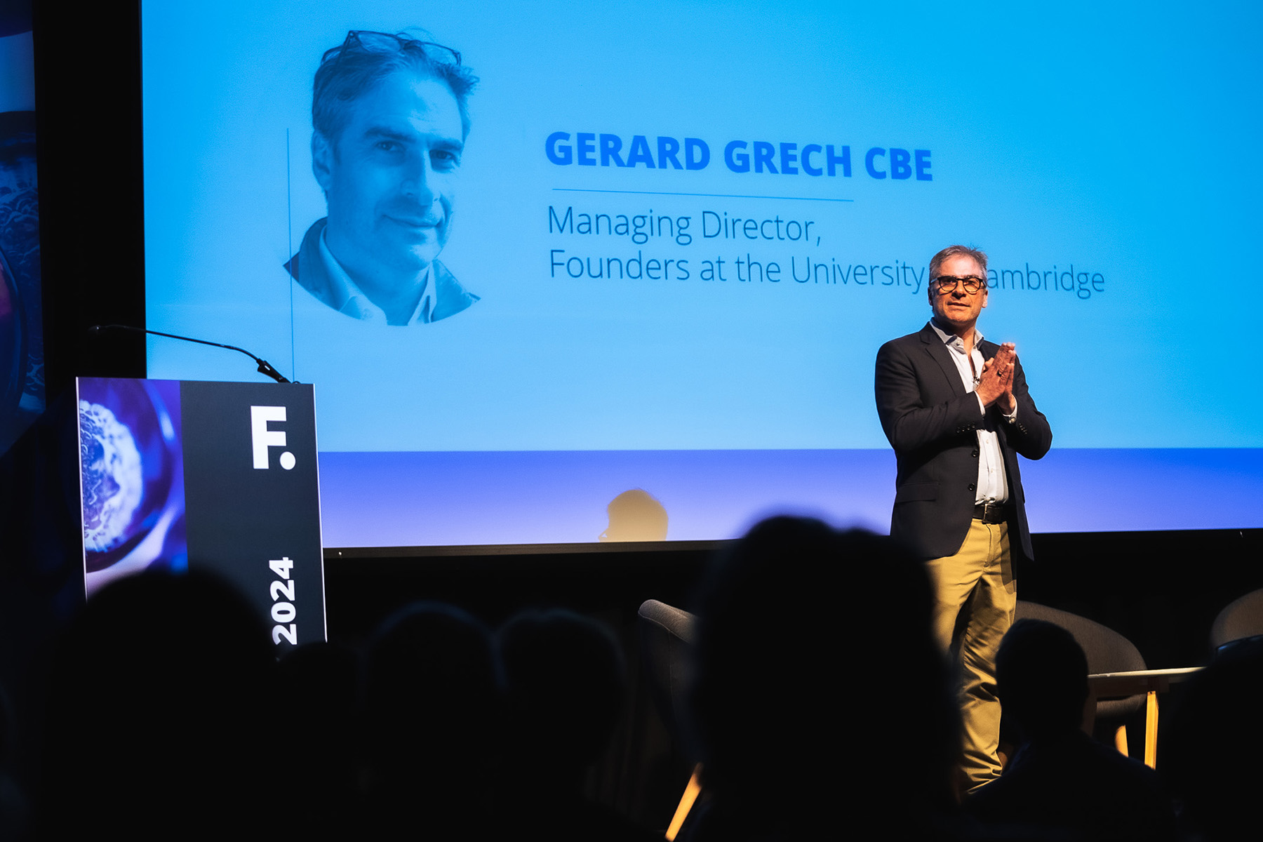 An individual standing on a stage, presenting to an audience with a large screen behind them. The screen displays the name “GERARD GRECH CBE” and the title “Managing Director, Founders at the University of Cambridge.”