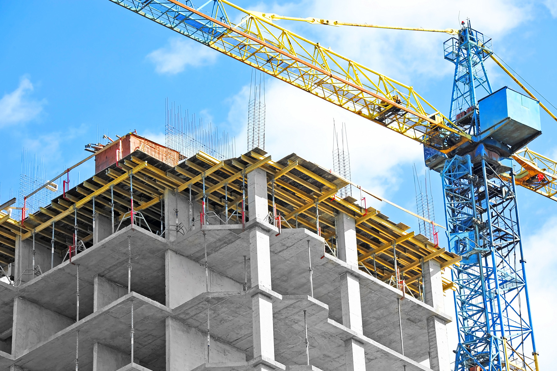 A construction site with a partially completed building and two tall cranes against a blue sky with clouds.
